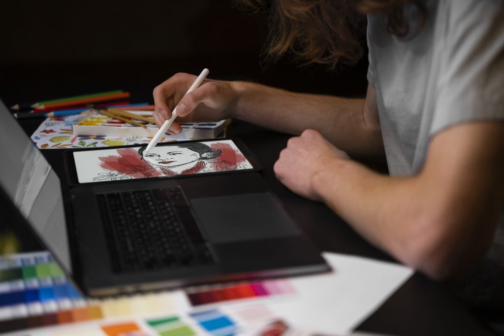 A man sitting at a desk with a color tray and printed images beside him, working on a laptop to design a book cover.