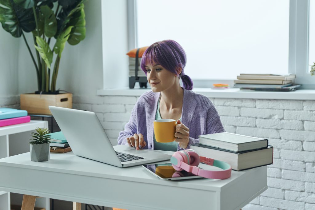 Woman with purple hair using a laptop at a white desk with books and a yellow coffee mug.