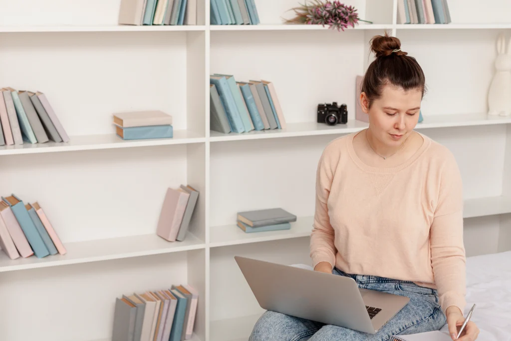 Woman sitting in front of white bookshelves using a laptop and taking notes for book writing.