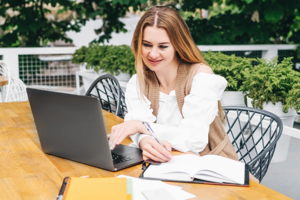 Author writing in a notebook next to a laptop on an outdoor wooden table surrounded by greenery.