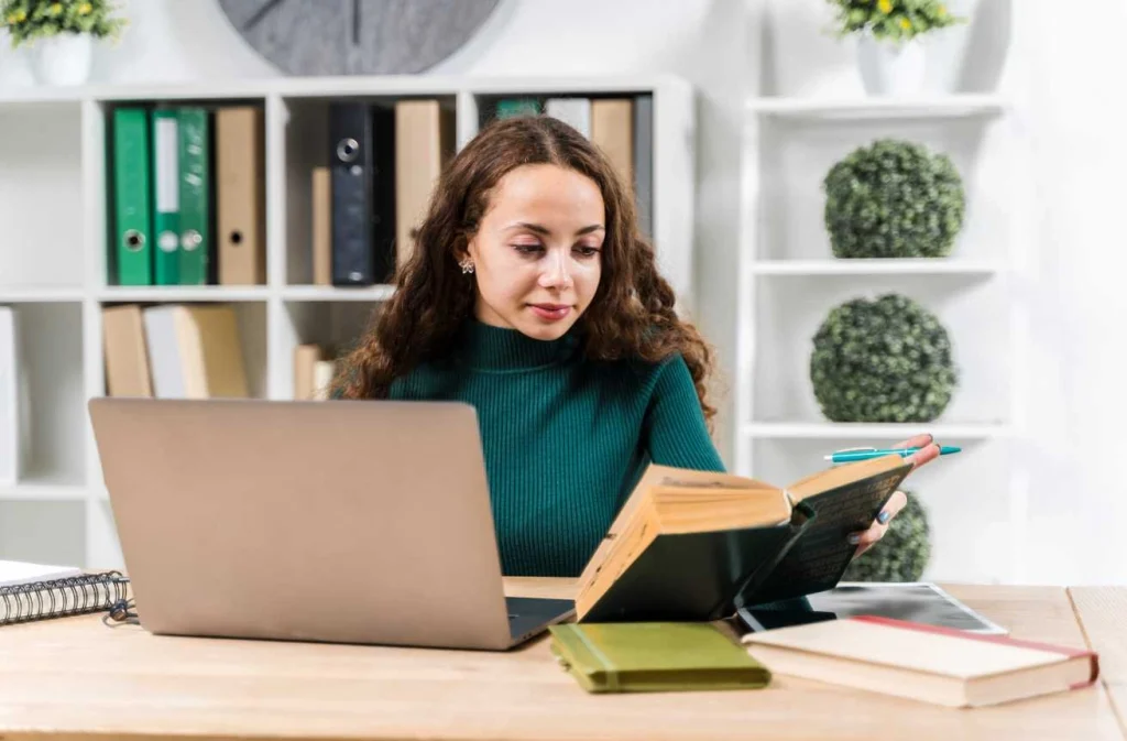 Woman reading book while using laptop at desk