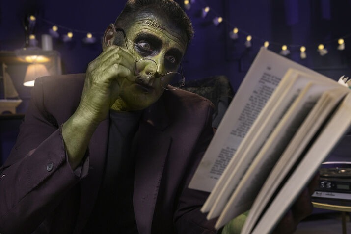 Person dressed as Frankenstein reading a horror book in a Halloween-themed setting.